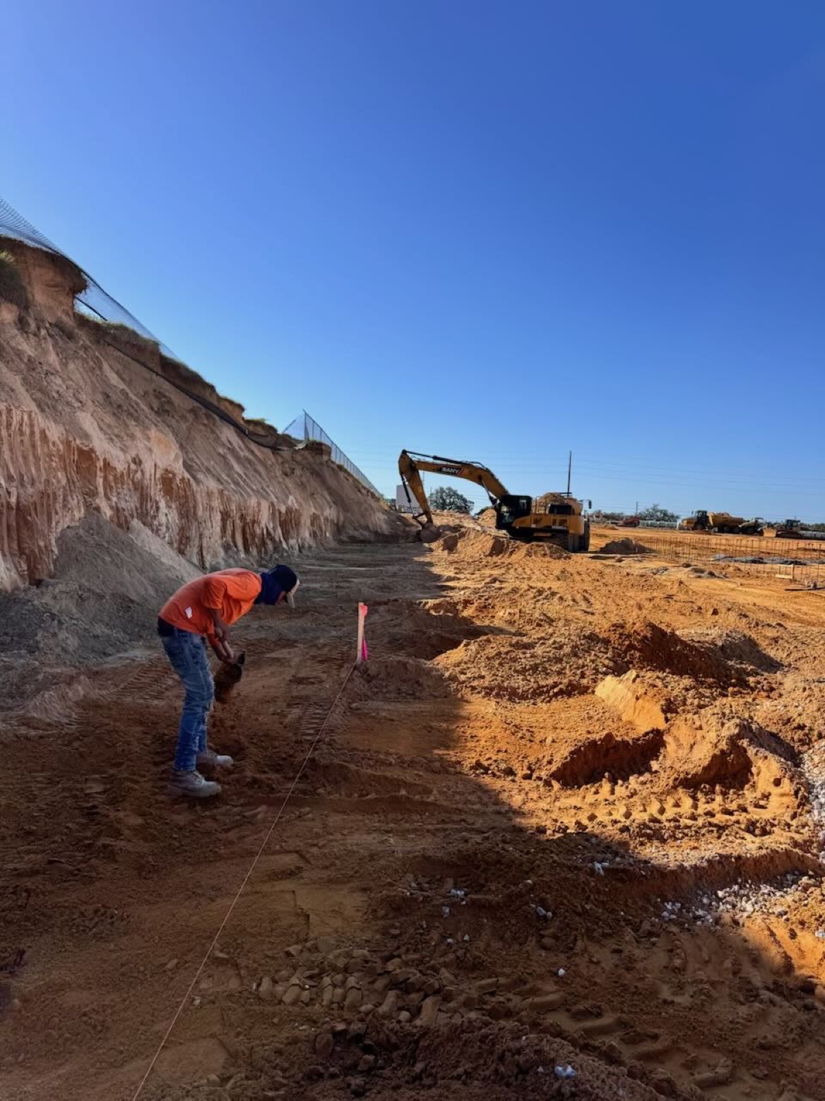 Crew member checking grade along a string line while an excavator works the cut behind him