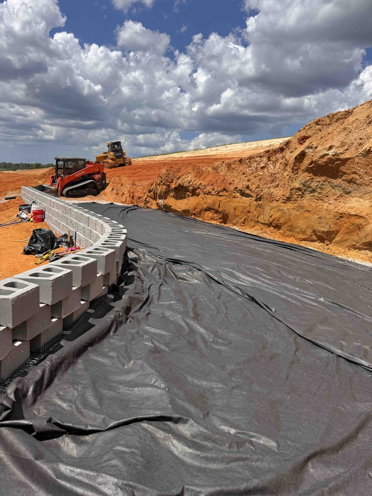 Block retaining wall going up beside a cut slope, with fabric laid behind it and a track loader working above